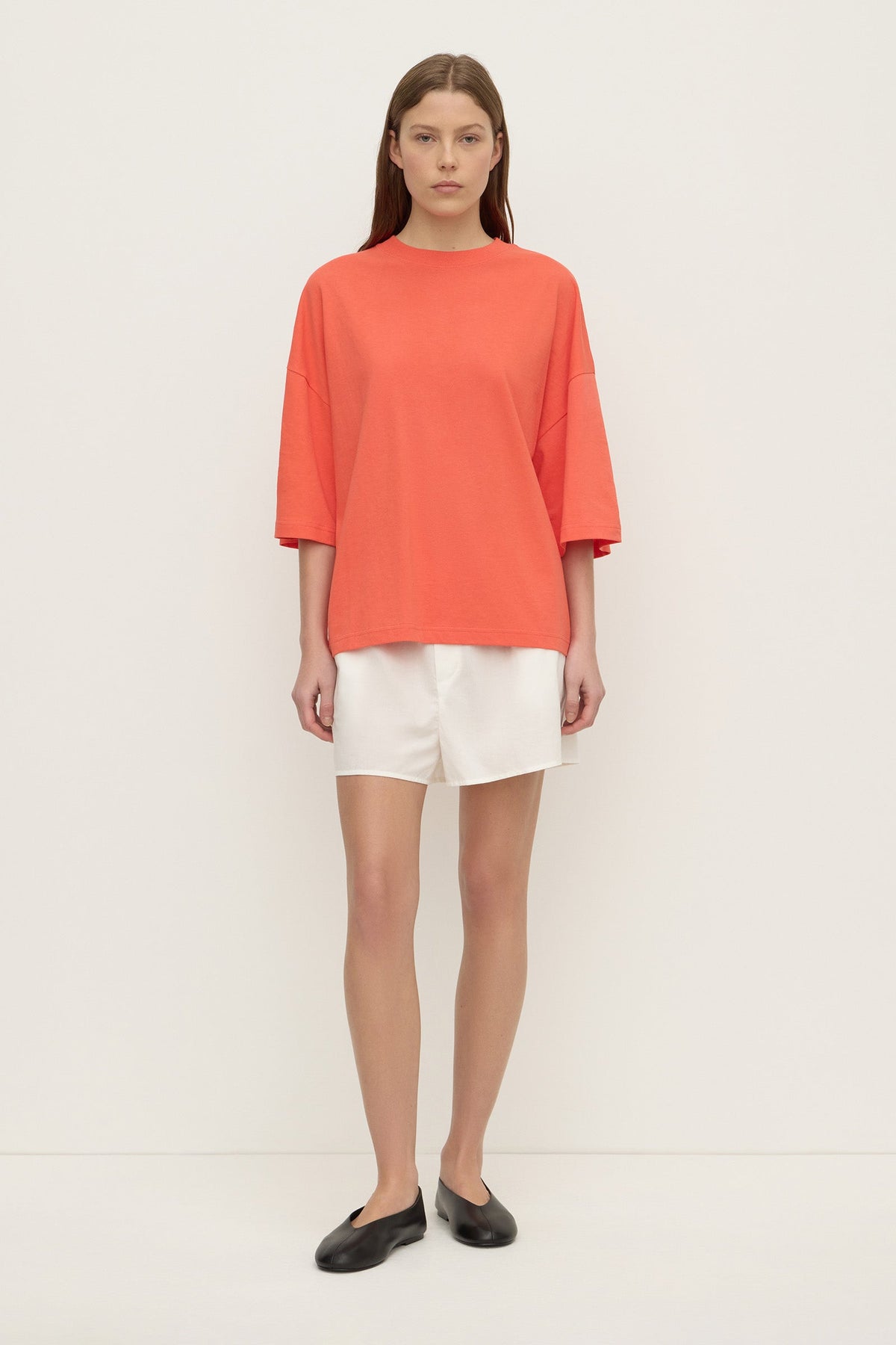 A model stands facing forward in a coral oversized T-shirt with a round neckline, relaxed body, dropped shoulders and wide elbow-length sleeves, paired with white shorts.