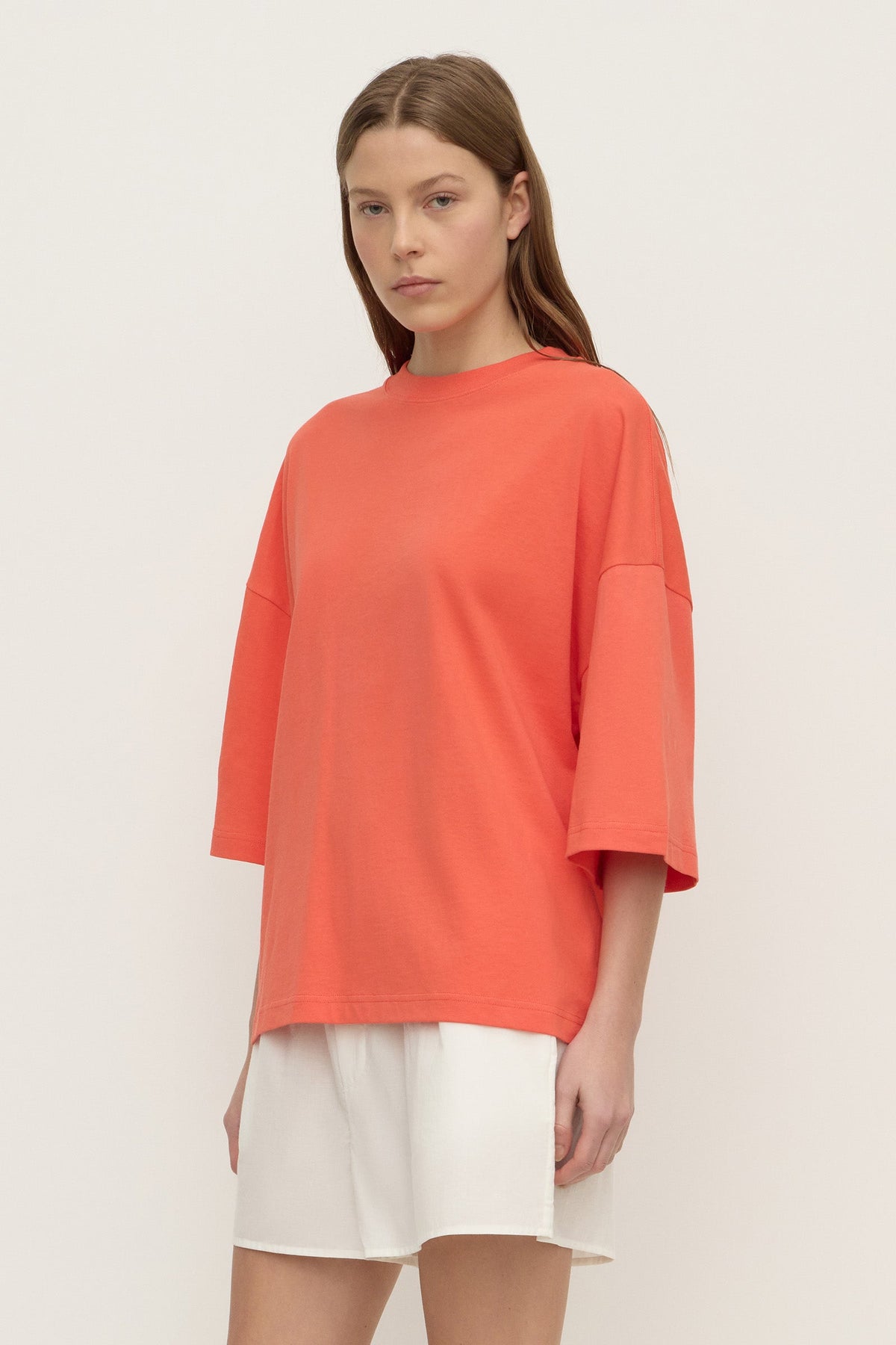 Three-quarter view of a model wearing a coral oversized T-shirt, showing the loose silhouette, dropped shoulder construction and wide elbow-length sleeves.