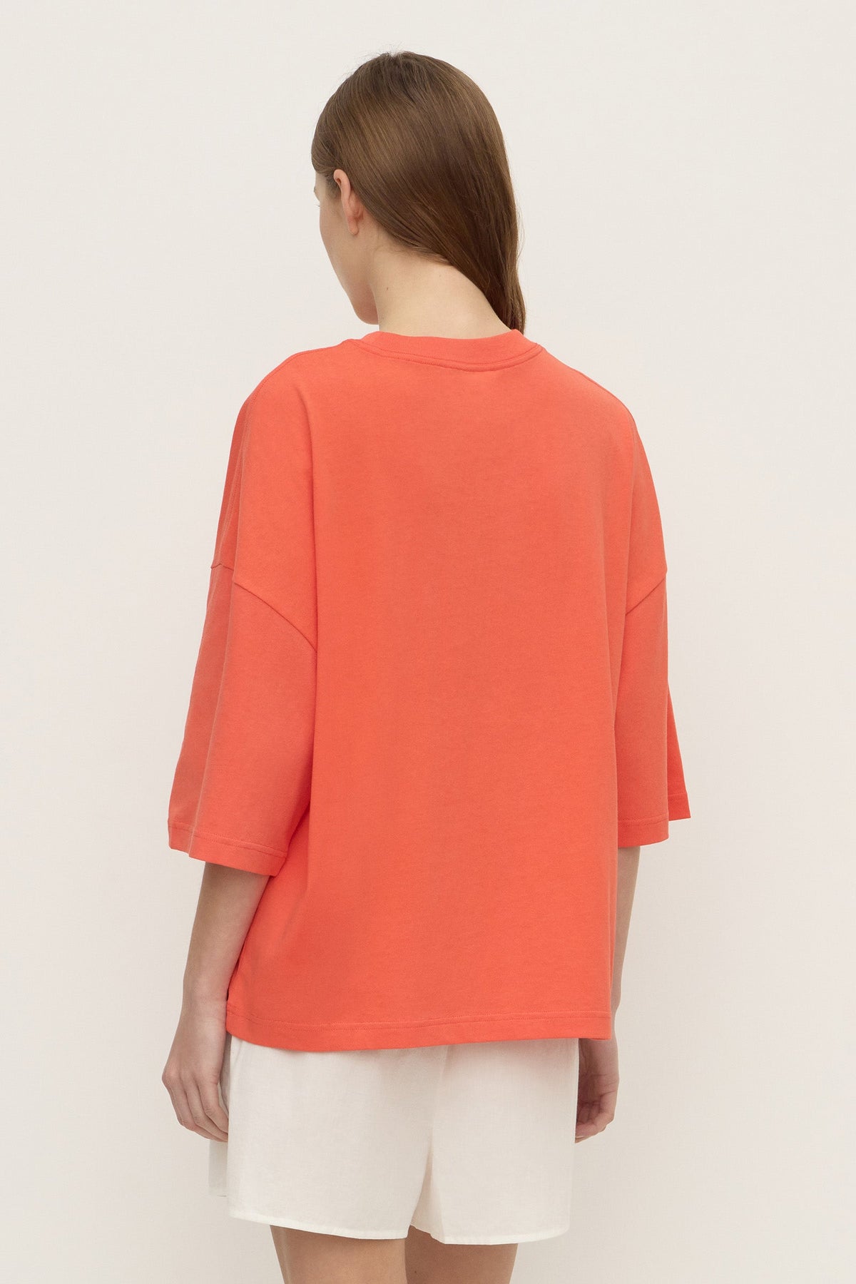 Back view of a model wearing a coral oversized T-shirt, clearly showing the round neckline, dropped shoulders, loose straight body and wide elbow-length sleeves.
