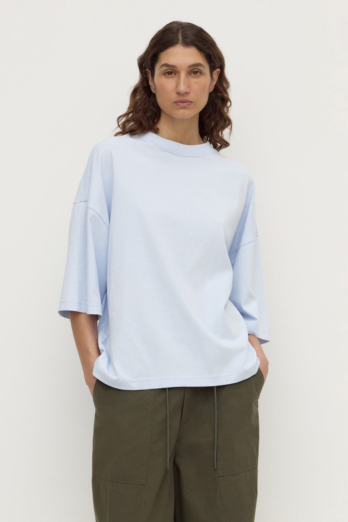 Model wearing a powder blue oversized short-sleeve tee with olive trousers, showing the crew neckline, dropped shoulders and relaxed fit, front view