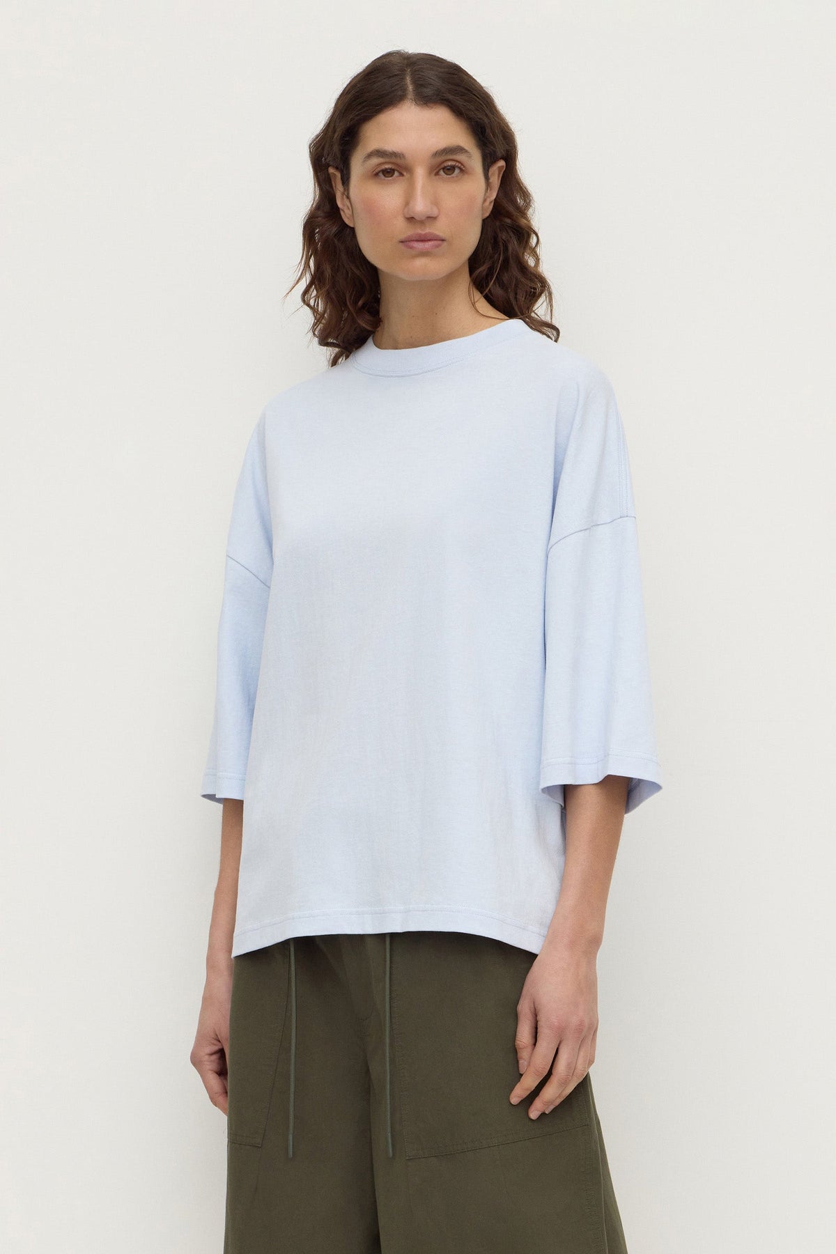 Model wearing a powder blue oversized short-sleeve tee with olive trousers, showing the crew neckline, dropped shoulders and straight hem, front view
