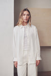 A woman modeling a white long-sleeve button-up shirt and matching white trousers in a minimalist indoor setting with neutral beige and gray walls.