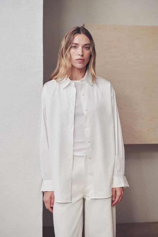 A woman modeling a white long-sleeve button-up shirt and matching white trousers in a minimalist indoor setting with neutral beige and gray walls.