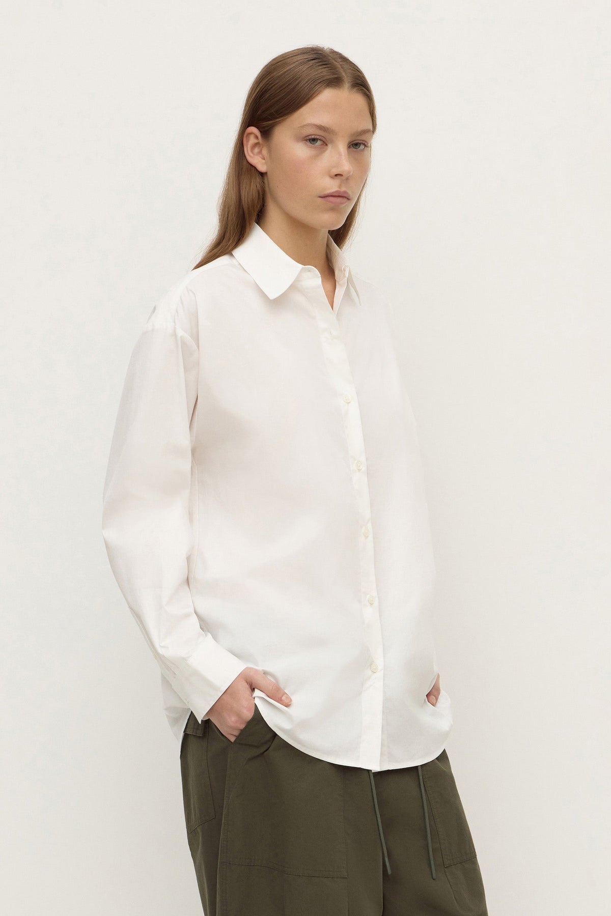 Model wearing a white long-sleeve button-up shirt with olive trousers, showing the curved hem and relaxed fit, angled front view