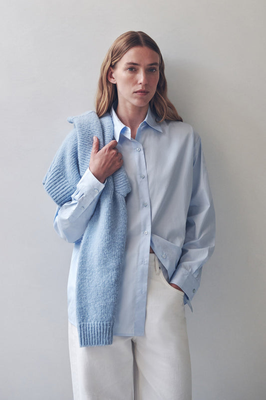 Model wearing a powder blue long-sleeve button-up shirt with white trousers and a coordinating knit draped over one shoulder, front view