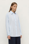 Model wearing a powder blue long-sleeve button-up shirt with navy trousers, showing the pointed collar, long sleeves and relaxed fit, angled front view
