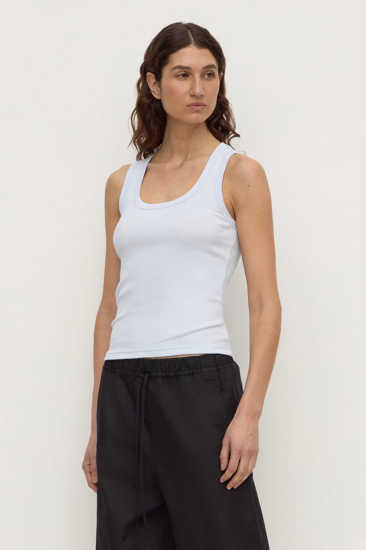 Model wearing a powder blue fitted rib tank with black trousers, showing the scoop neckline and close-fitting silhouette, angled front view
