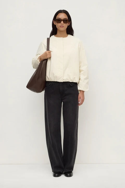 A model stands against a plain white studio background wearing a cream-colored long-sleeve top, black wide-leg trousers, black shoes, sunglasses, and carrying a brown leather shoulder bag.