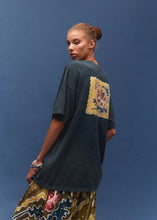 A woman wearing an oversized dark charcoal t-shirt with a large yellow and blue floral patch on the back, paired with a patterned skirt, set against a solid blue studio background.