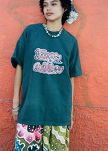 A person wearing an oversized dark teal graphic T-shirt with a pink and white bubble-letter design reading 'Kings of NY', paired with colorful patterned pants, standing against a textured orange wall.