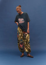 A person stands against a blue studio backdrop wearing a dark oversized graphic T-shirt and loose patterned harem-style pants with brown studded sandals.
