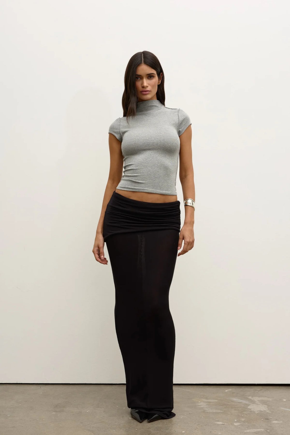 Model wearing a Grey Marle fitted cap-sleeve mock-neck top with a black ruched maxi skirt and pointed shoes, showing the cropped length and slim silhouette, front view