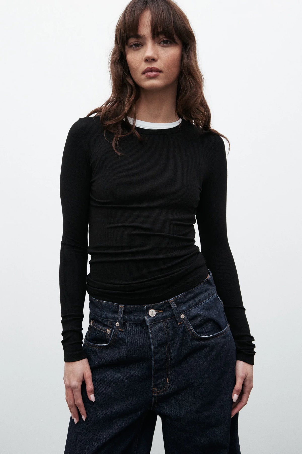 Woman wearing a black long-sleeve top and dark jeans against a white background