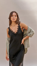 Woman wearing a black slip dress with lace detailing and an olive green jacket on a neutral background
