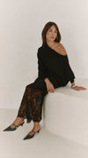 A model seated in the black skirt, showing its asymmetrical overlay and floral lace detailing styled with a black off-shoulder top.
