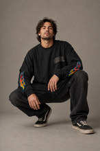 Man crouching in the oversized black long sleeve tee, highlighting the outlined chest logo and colourful flame graphics along both sleeves against a neutral studio backdrop.