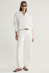 A model wearing a white cropped button-up shirt with a pointed collar and long sleeves, paired with white straight-leg trousers, a black belt and black flat sandals in a full-length front view.