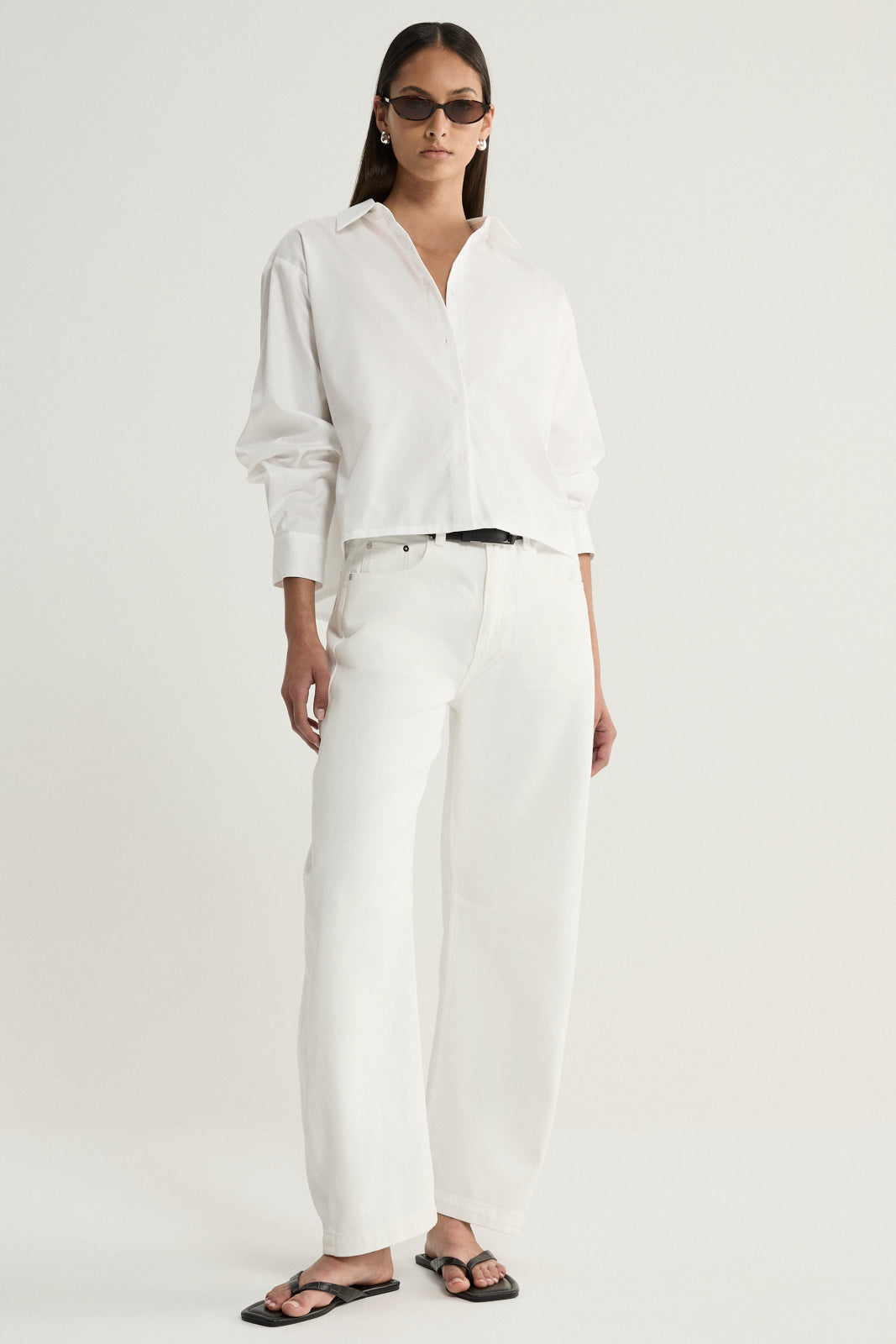 A model wearing a white cropped button-up shirt with a pointed collar and long sleeves, paired with white straight-leg trousers, a black belt and black flat sandals in a full-length front view.