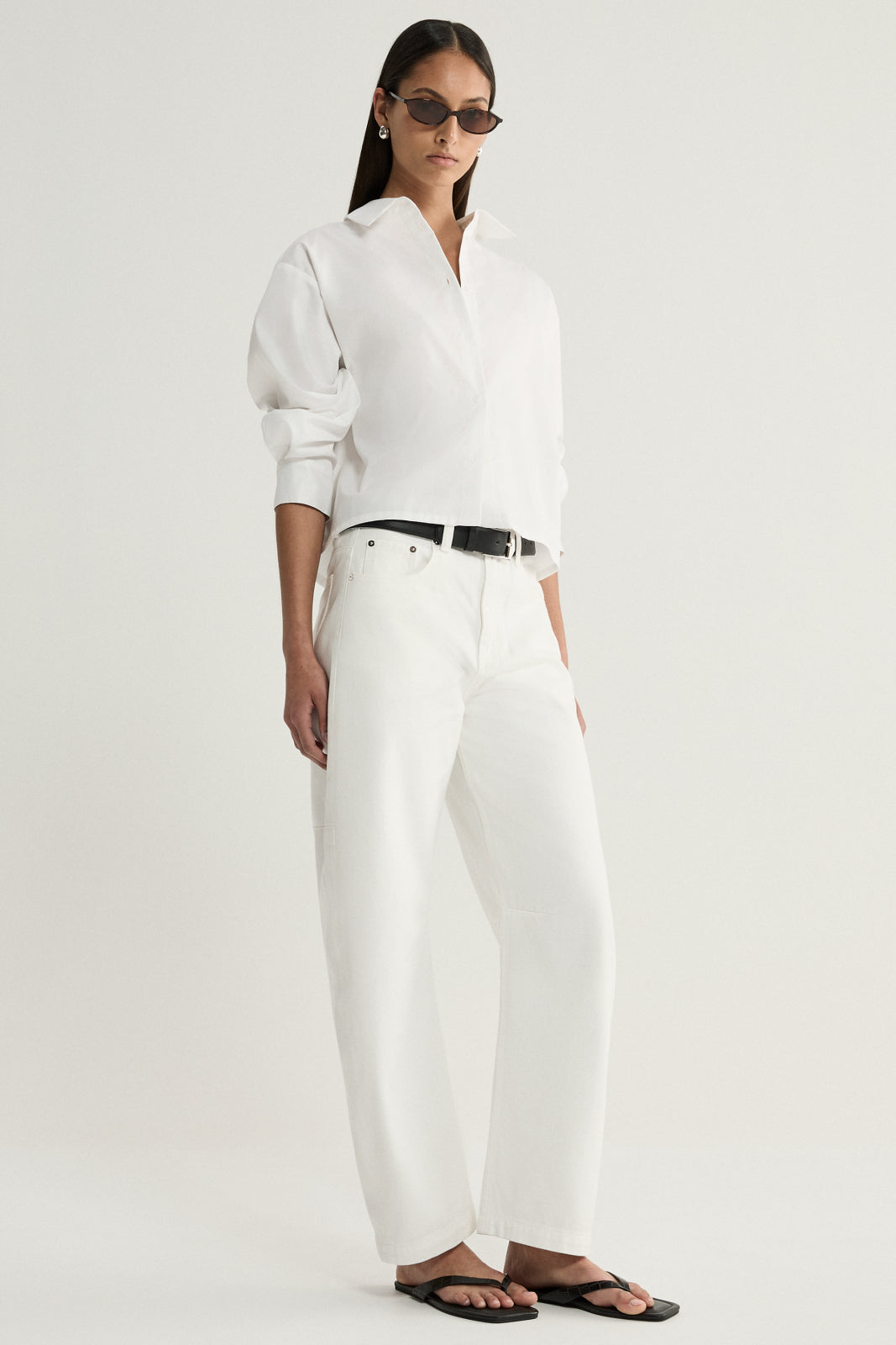A three-quarter view of a model wearing a white cropped button-up shirt with a pointed collar, relaxed long sleeves and button cuffs, paired with white trousers and a black belt.