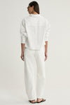 A rear view of a model wearing a white cropped shirt showing the back panel, shoulder seams and relaxed sleeves, paired with white trousers and black flat sandals.