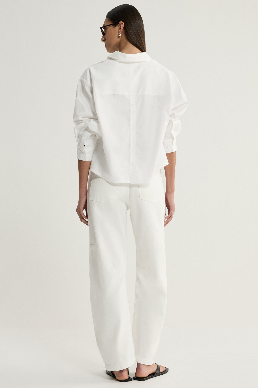 A rear view of a model wearing a white cropped shirt showing the back panel, shoulder seams and relaxed sleeves, paired with white trousers and black flat sandals.