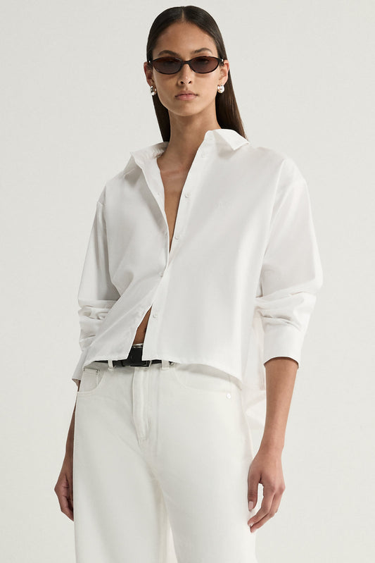 A close-up of a white cropped shirt showing its pointed collar, front button placket, open neckline and long sleeve with an adjustable cuff.