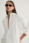 A close-up of a model wearing a white cropped button-up shirt with a wide open collar, visible front buttons and relaxed sleeves, showing the crisp fabric and loose shape.