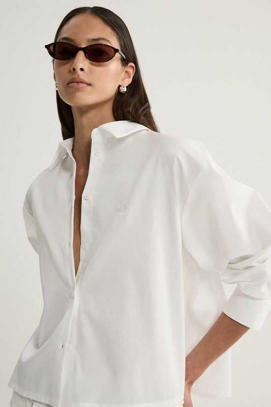 A close-up of a model wearing a white cropped button-up shirt with a wide open collar, visible front buttons and relaxed sleeves, showing the crisp fabric and loose shape.