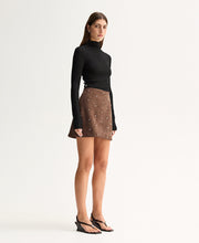 Model wearing the Mocha Elka Collective Avenue Mini Skirt with a black turtleneck knit top, showing the high-waisted fit and subtly flared hem, full-length angled view
