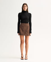 Model wearing the Mocha Elka Collective Avenue Mini Skirt with a black turtleneck knit top, showing the A-line silhouette and all-over silver-tone dome studs, full-length front view