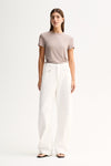A model wearing an almond-grey fitted short-sleeve tee with a round neckline, tucked into white wide-leg trousers and paired with black flat sandals in a full-length front view.