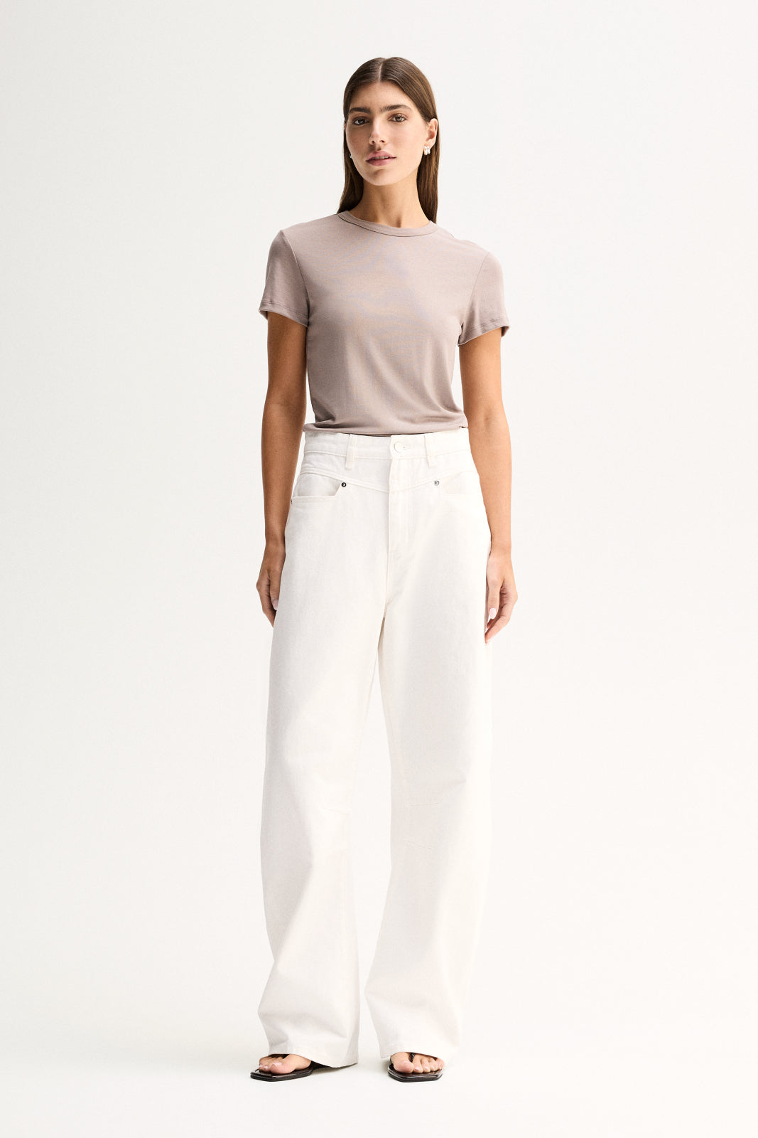 A model wearing an almond-grey fitted short-sleeve tee with a round neckline, tucked into white wide-leg trousers and paired with black flat sandals in a full-length front view.
