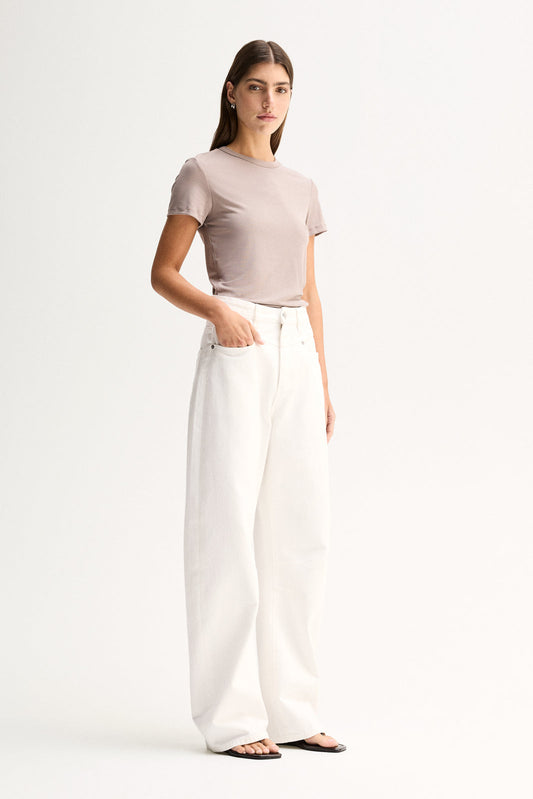 A model wearing an almond-grey fitted tee with short sleeves and a round neckline, styled with white wide-leg trousers and black sandals in an angled front pose.