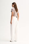 A rear three-quarter view of a model wearing an almond-grey short-sleeve tee with a simple back and regular-length fit, paired with white wide-leg trousers.
