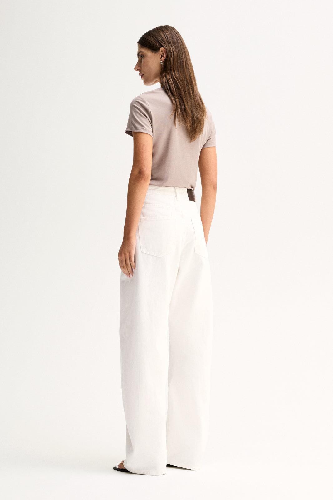 A rear three-quarter view of a model wearing an almond-grey short-sleeve tee with a simple back and regular-length fit, paired with white wide-leg trousers.