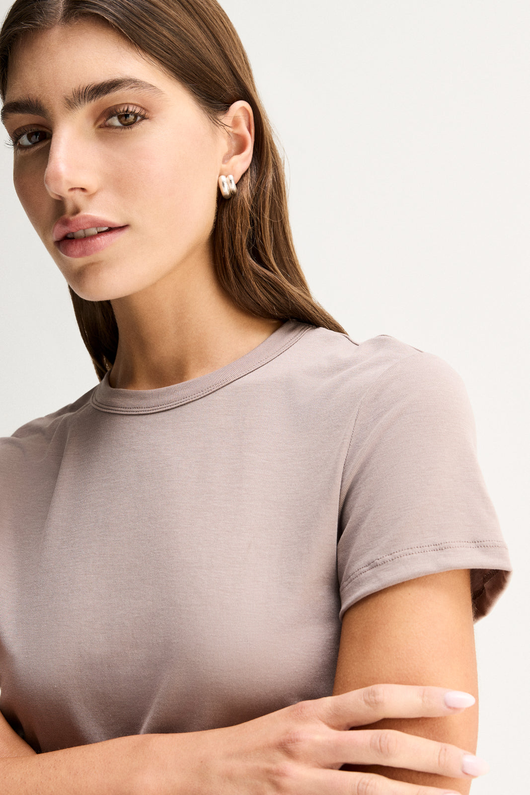 A close-up of an almond-grey fitted tee showing its round bound neckline, short sleeve and smooth fine-textured fabric, with the model's arms folded across the front.