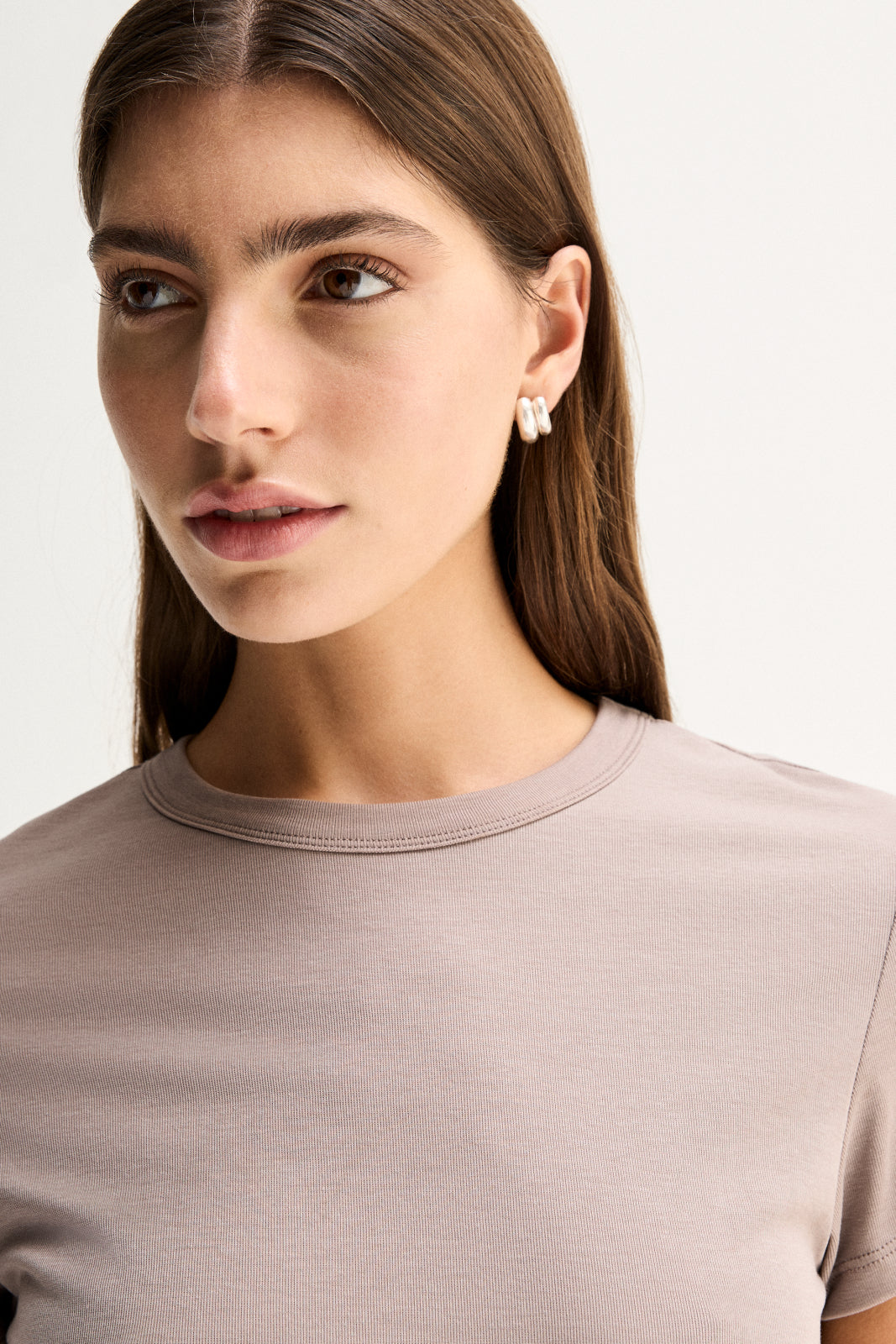 A close-up of an almond-grey fitted tee showing its round bound neckline, short sleeve and smooth fine-textured fabric, with the model's arms folded across the front.