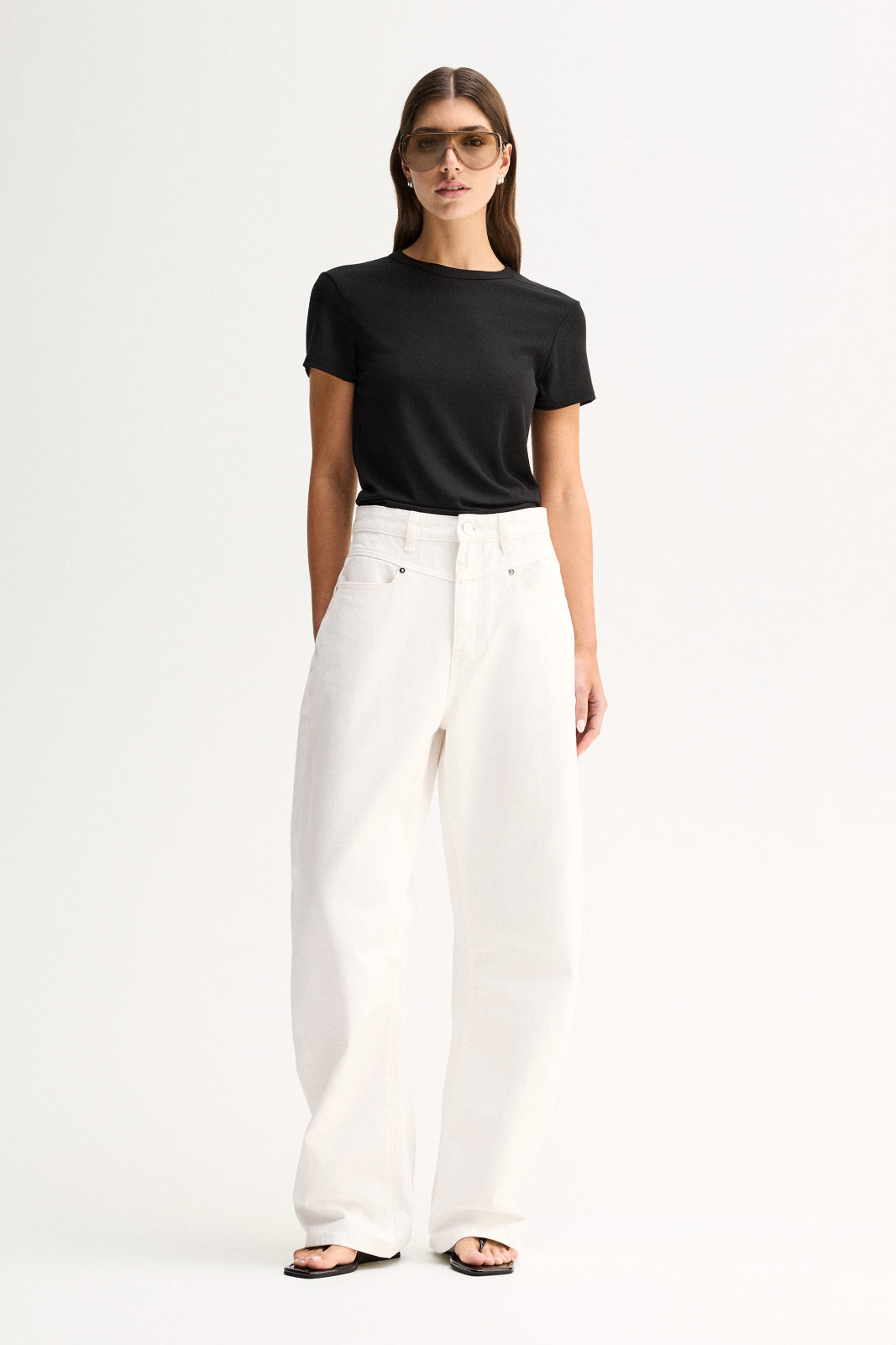 A model wearing a fitted black short-sleeve tee with a round neckline, tucked into white wide-leg trousers and paired with flat black sandals in a full-length front view.