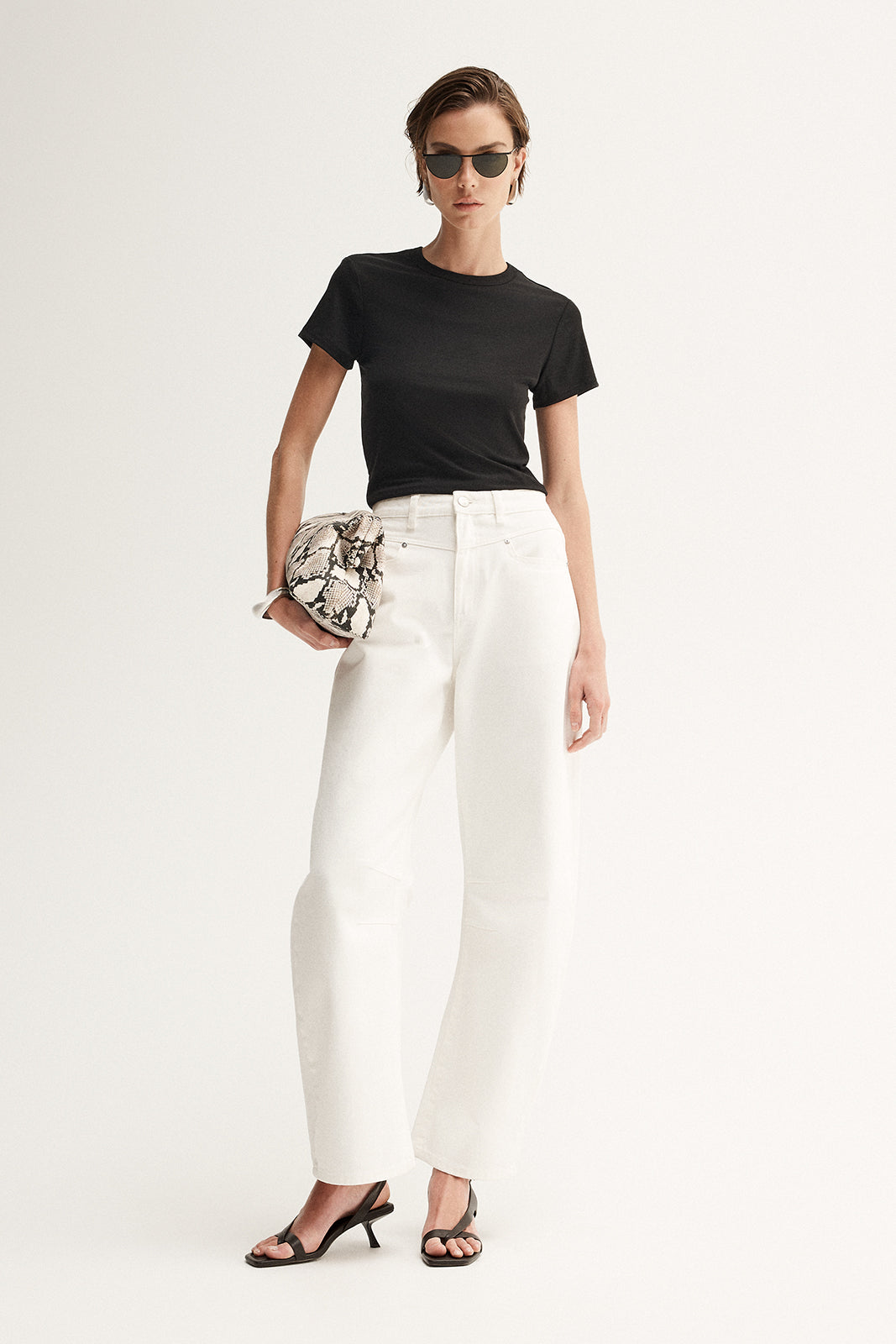 A model wearing a fitted black crew-neck tee and white wide-leg trousers, accessorised with dark sunglasses, a patterned clutch and black heeled sandals in a front-facing studio pose.