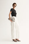 A model in a fitted black short-sleeve tee and white wide-leg trousers holds a cream-and-black patterned clutch, shown from an angled side view with black strappy heels.