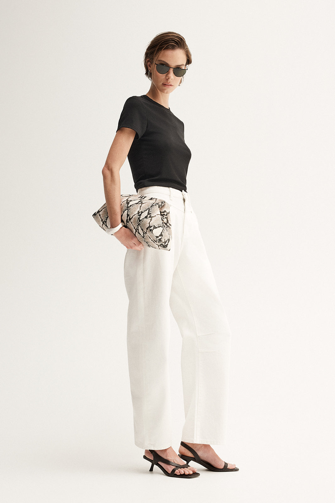 A model in a fitted black short-sleeve tee and white wide-leg trousers holds a cream-and-black patterned clutch, shown from an angled side view with black strappy heels.