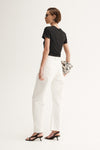 A rear three-quarter view of a model wearing a black short-sleeve tee with a simple back and white wide-leg trousers, carrying a patterned clutch and wearing black heeled sandals.