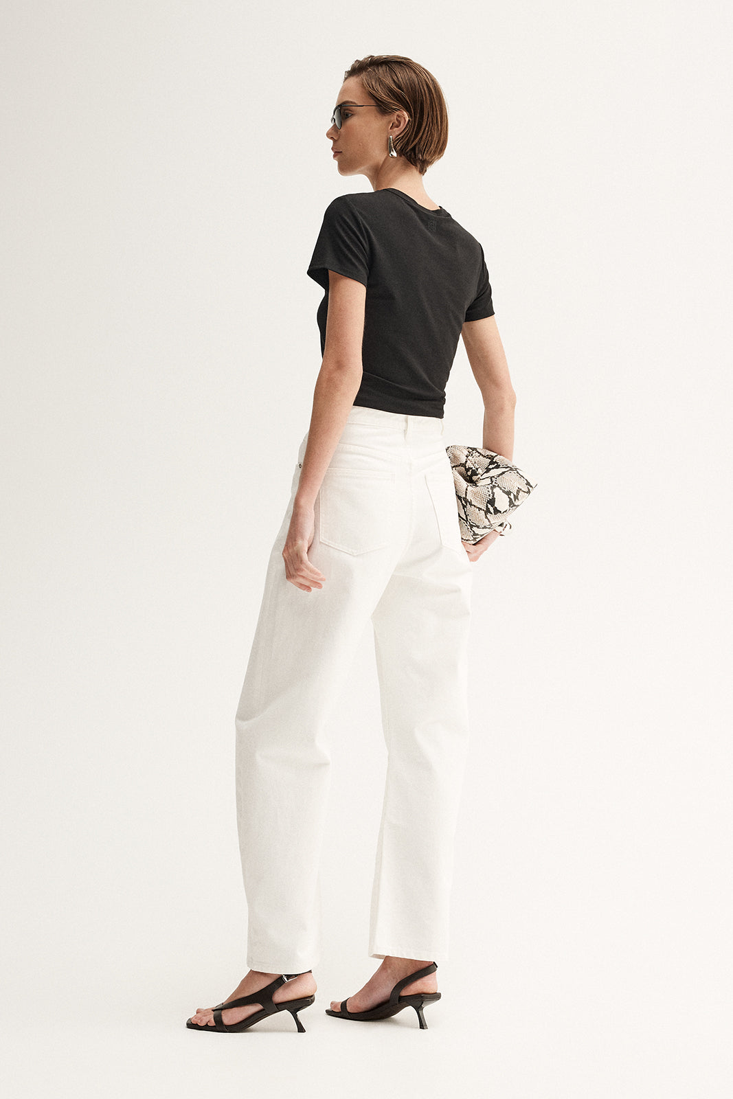 A rear three-quarter view of a model wearing a black short-sleeve tee with a simple back and white wide-leg trousers, carrying a patterned clutch and wearing black heeled sandals.
