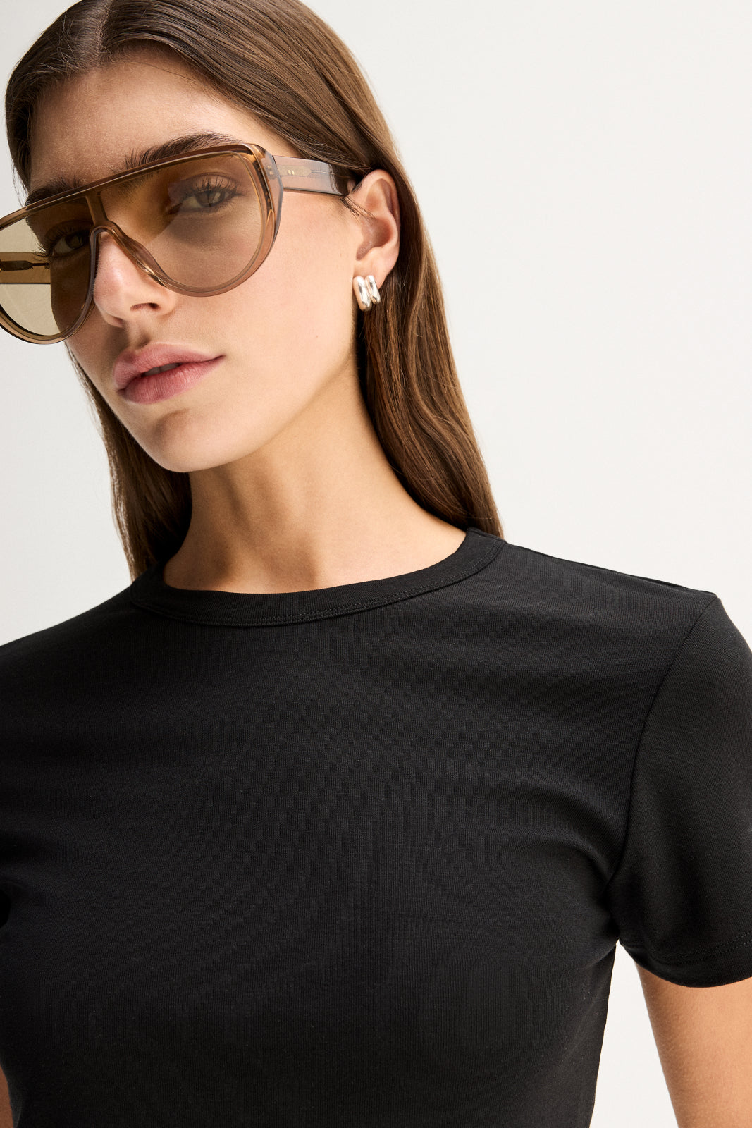 A close-up of a black fitted tee showing its round bound neckline, short sleeve and smooth fabric, styled with oversized brown sunglasses and small silver earrings.