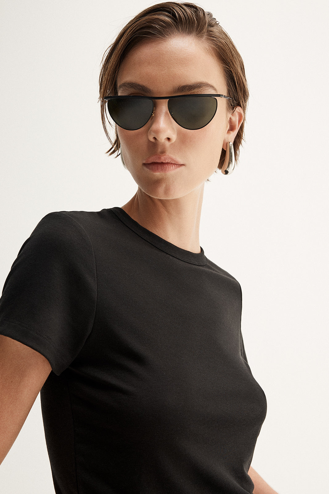 A close-up of a model wearing a black fitted tee with a classic round neckline and short sleeves, showing the clean collar finish and close-fitting shape alongside dark sunglasses and silver hoop earrings.