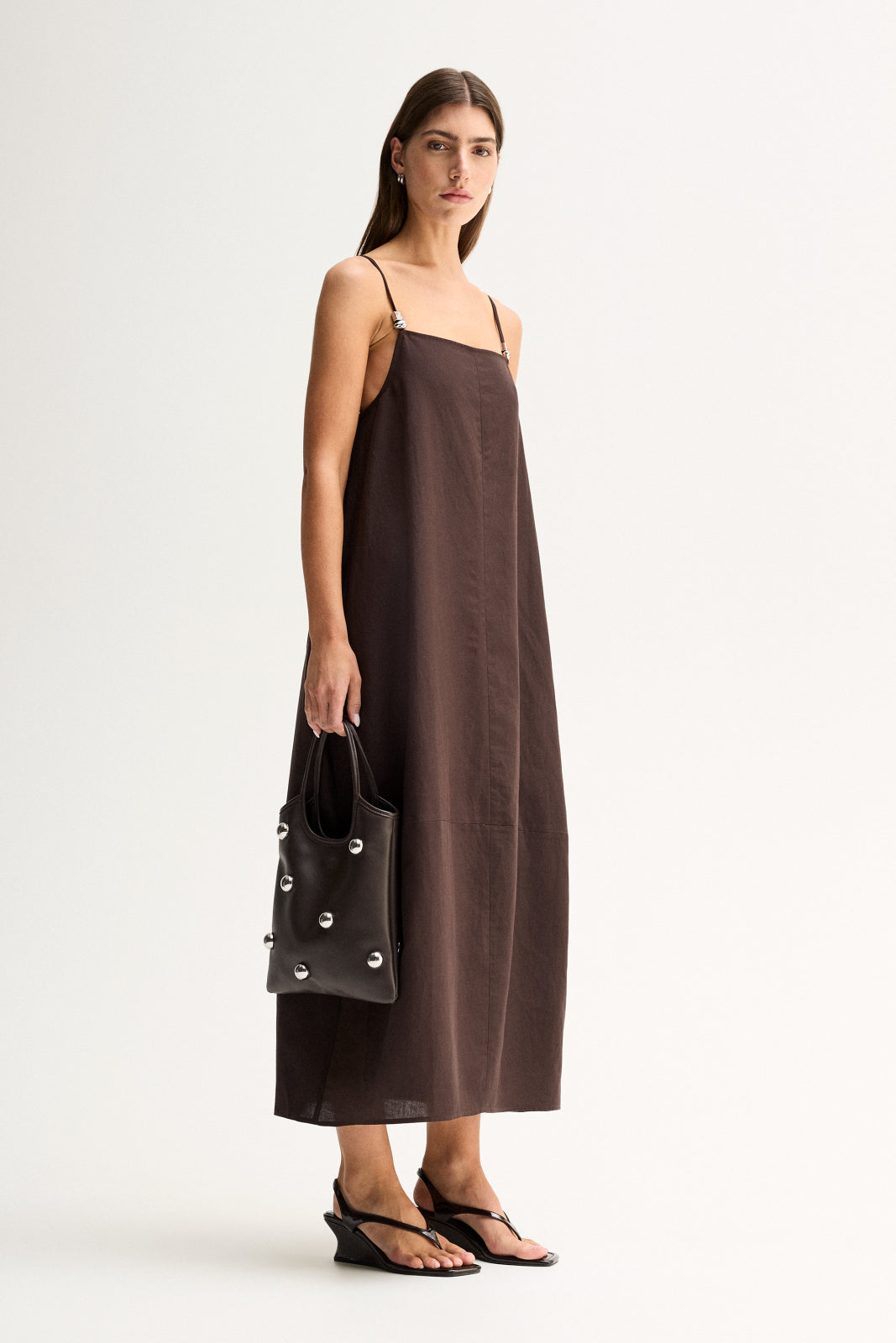 A woman wearing a chocolate maxi dress with thin adjustable straps and a relaxed silhouette, carrying a black handbag.