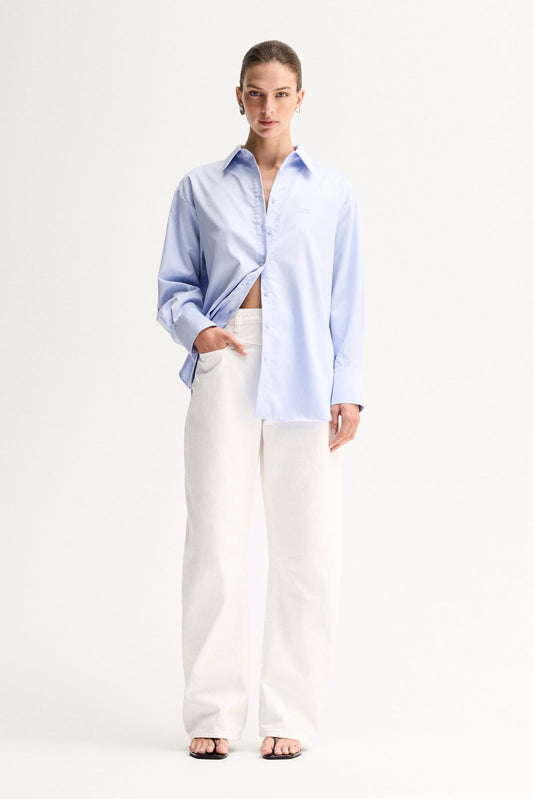 Model wearing the Light Blue Elka Collective Carter Shirt with white wide-leg jeans, showing the oversized silhouette and relaxed drape falling open at the side, full-length angled view