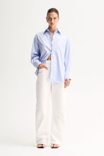 Model wearing the Light Blue Elka Collective Carter Shirt with white wide-leg jeans, showing the oversized silhouette and relaxed drape falling open at the side, full-length angled view