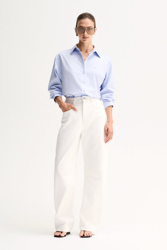 Model wearing the Light Blue Elka Collective Carter Shirt with white wide-leg jeans, black thong sandals and sunglasses, showing the relaxed oversized fit, spread collar and button-through front, full-length front view
