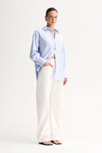 Model wearing the Light Blue Elka Collective Carter Shirt with white wide-leg jeans and black thong sandals, showing the button-through front and long sleeves with turned-back cuffs, full-length side view